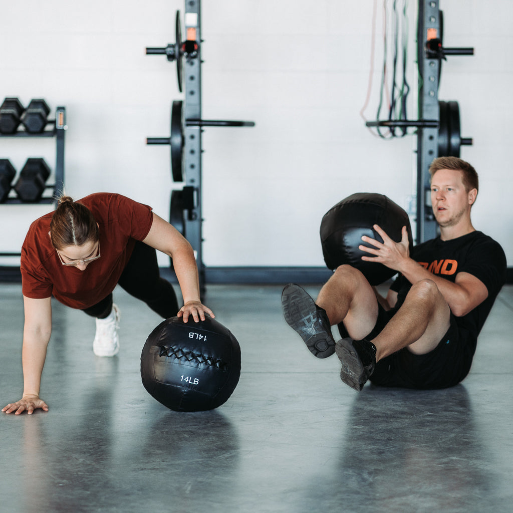 Two people working out with Soft Wall Balls. One doing Russian twists and one using Soft Wall Ball for one arm elevated push up.