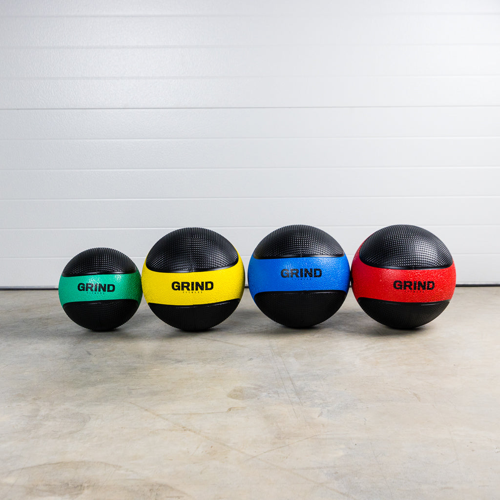 Green, Yellow, Blue, and Red GRIND Medicine Balls in a row on the floor. Each color depicts a different weight. 